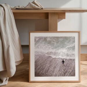 Könnte beinhalten: Ein gerahmter Druck, der eine Person zeigt, die ins Meer geht. Das Bild fängt den Moment ein, in dem die Person ins Wasser geht, mit Wellen, die an die Küste schlagen. Der Rahmen ist helles Holz, und der Druck hat eine neutrale Farbpalette.