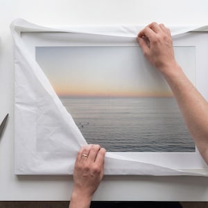 Könnte beinhalten: Ein rechteckiger gerahmter Druck mit einer Meereslandschaft bei Sonnenuntergang, einem sanften Himmelsverlauf und ruhigen Wellen, teilweise in weißes Seidenpapier eingewickelt. Ein Abroller und eine Schere liegen neben dem Rahmen.