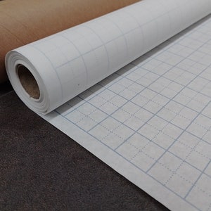 May include: A roll of white grid paper with light blue lines, partially unrolled, next to a brown cardboard tube. The paper is on a dark brown surface. The grid pattern is visible on the unrolled portion.