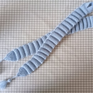 May include: A light blue knitted scarf with two tassels. The scarf is made of a soft, textured yarn and has a simple, elegant design.