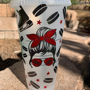 May include: A white reusable cup with a frosted finish features a graphic design of a woman with a messy bun, red sunglasses, and a red bow. The design also includes hockey pucks and red stars.