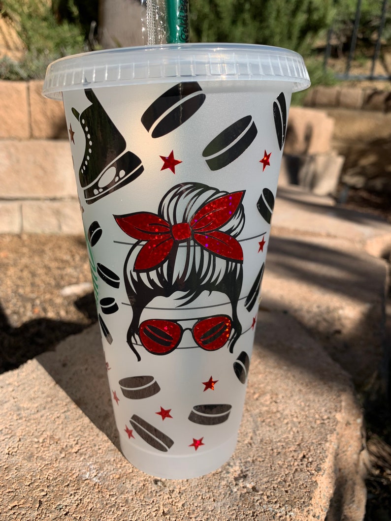 May include: A white reusable cup with a frosted finish features a graphic design of a woman with a messy bun, red sunglasses, and a red bow. The design also includes hockey pucks and red stars.
