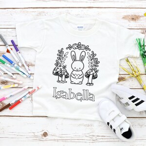Pode incluir: T-shirt branca de manga curta com um desenho de coelho e cogumelos a preto e branco, e o nome "Isabella". Inclui um par de ténis brancos e marcadores coloridos. É uma peça de vestuário infantil.