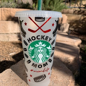 May include: A white reusable cup with a black and green Starbucks logo and the words "Hockey Mom #momlife" in red. The cup is decorated with hockey sticks, pucks, and stars.