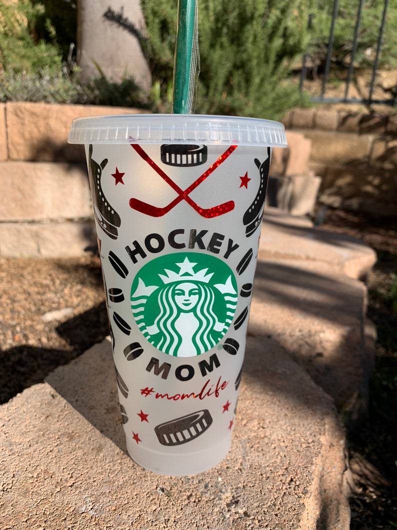 May include: A white reusable cup with a black and green Starbucks logo and the words "Hockey Mom #momlife" in red. The cup is decorated with hockey sticks, pucks, and stars.