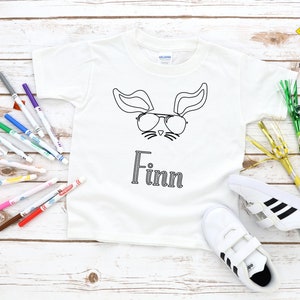 Pode incluir: Camiseta branca com um contorno preto de um coelho de óculos de sol e o nome "Finn" embaixo. A camiseta está cercada por marcadores e suprimentos para festas.