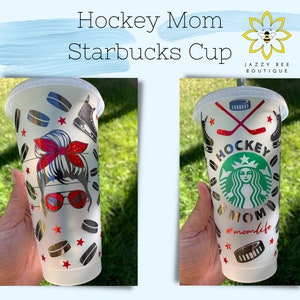 May include: Two white Starbucks cups with a hockey theme. One cup features a graphic of a woman with a red headband and sunglasses, and the other cup features a graphic of the Starbucks logo with the words "Hockey Mom" and "#momlife". Both cups have a black and silver hockey puck pattern.