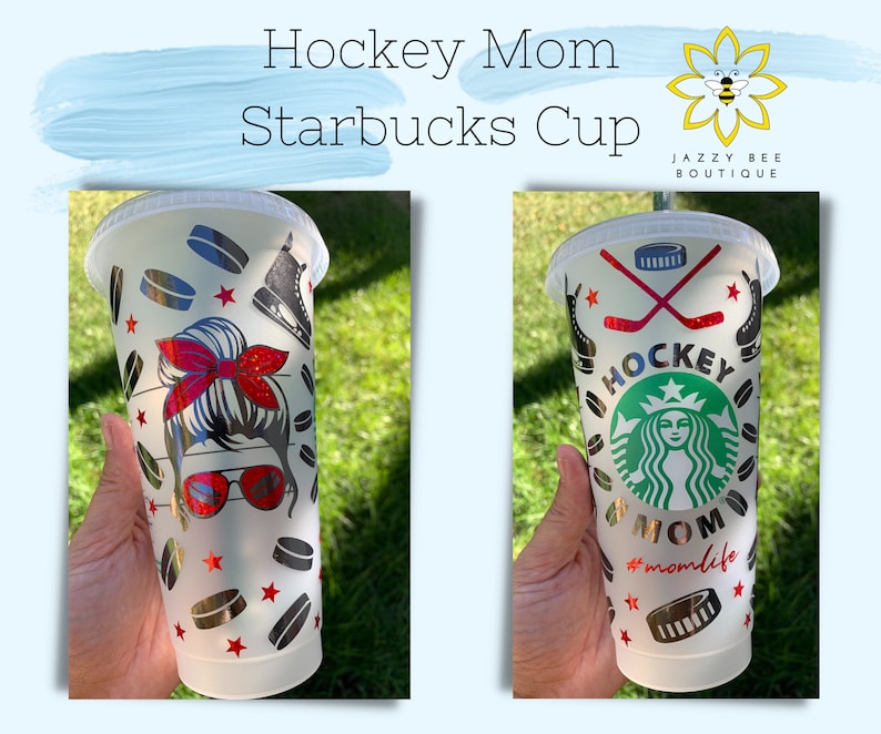May include: Two white Starbucks cups with a hockey theme. One cup features a graphic of a woman with a red headband and sunglasses, and the other cup features a graphic of the Starbucks logo with the words "Hockey Mom" and "#momlife". Both cups have a black and silver hockey puck pattern.