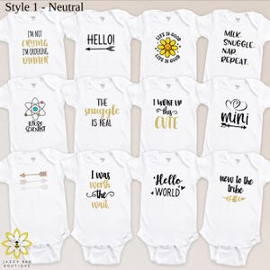 Pu&ograve; includere: Una collezione di body bianchi per neonati con vari disegni di testo neri e oro. Le frasi includono "I'm not crying, I'm ordering dinner", "Hello!" e "Life is Good". I body sono di Jazzy Bee Boutique.