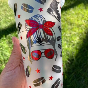 May include: A white reusable cup featuring a design of a woman with a red bow in her hair, wearing sunglasses and a hockey skate. The design also includes hockey pucks and red stars.