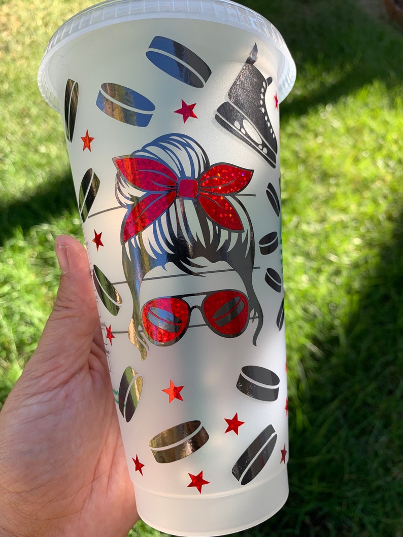 May include: A white reusable cup featuring a design of a woman with a red bow in her hair, wearing sunglasses and a hockey skate. The design also includes hockey pucks and red stars.