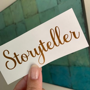 Pode incluir: Um cart&atilde;o branco com a palavra "Storyteller" escrita em letras douradas em it&aacute;lico.