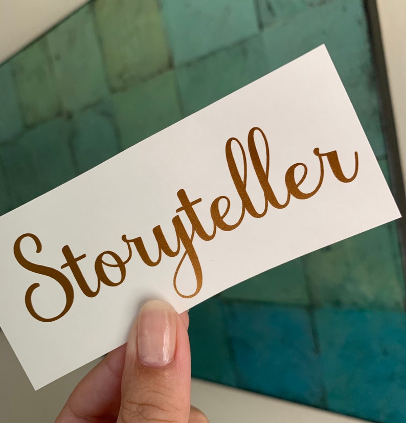 Pode incluir: Um cart&atilde;o branco com a palavra "Storyteller" escrita em letras douradas em it&aacute;lico.
