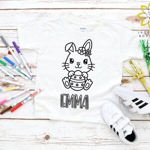 Pode incluir: Camiseta branca com um contorno preto e branco de um coelho segurando um ovo de Páscoa. O coelho tem uma coroa de flores e o nome "EMMA" está escrito abaixo do coelho. A camiseta está cercada por marcadores e outros materiais de artesanato.