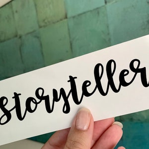 Pode incluir: Cart&atilde;o branco com a palavra "storyteller" escrita em caligrafia preta.