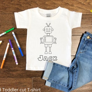 May include: White toddler t-shirt with a black and white outline of a robot and the name "JACK" underneath. The shirt is laying on a wooden surface with colored markers and blue jeans.