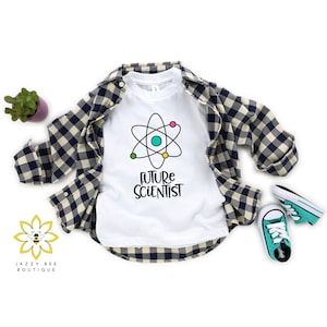 May include: White t-shirt featuring a black atom graphic and the text "FUTURE SCIENTIST". The shirt is laid out on a checkered fabric with a pair of teal trainers.