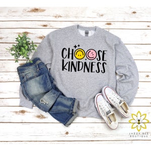 May include: Gray sweatshirt with the text "Choose Kindness" in black lettering. Two smiley faces, one yellow and one pink, are above the text. The sweatshirt is laid out on a wood surface with a pair of white sneakers and blue jeans.