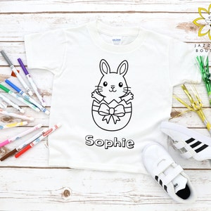 Pode incluir: Camiseta branca com um contorno preto e branco de um coelho dentro de um ovo. O coelho tem um laço na cabeça. A camiseta diz "Sophie" abaixo da imagem. A camiseta está cercada por marcadores e artigos de festa.