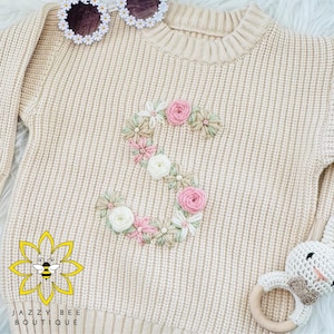 May include: A beige knitted sweater with a floral embroidered letter 'S' on the front. The sweater is made of a soft, comfortable material and has a ribbed texture.
