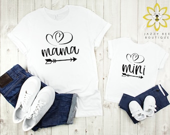 Könnte beinhalten: Zwei weiße T-Shirts mit schwarzem Text und Grafiken. Das größere Shirt sagt "mama" mit einem Herzen und einem Pfeil. Das kleinere Shirt sagt "mini" mit einem Herzen und einem Pfeil.