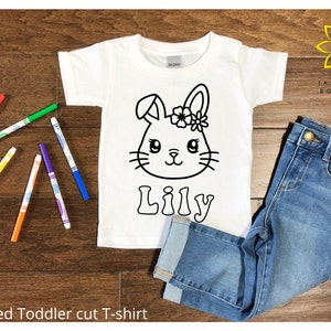 Pode incluir: Camiseta branca para criança pequena com um contorno preto e branco de um coelho com flores na cabeça. O nome "Lily" está escrito abaixo do coelho. A camiseta está sobre uma superfície de madeira com marcadores coloridos.