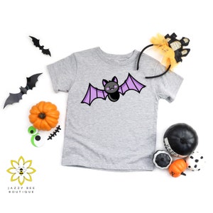 May include: A grey t-shirt featuring a purple bat graphic. The bat has black eyes and a small mouth. The shirt is on a white background with Halloween decorations.