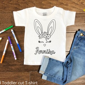 Pode incluir: Camiseta branca para criança pequena com um contorno preto de um coelho usando uma coroa de flores. O nome "Annika" está escrito abaixo do coelho. A camiseta é mostrada com marcadores coloridos e calças jeans azuis.
