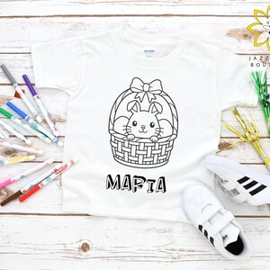 Pode incluir: Camiseta branca com um desenho a preto e branco de um coelho numa cesta, com o nome "Maria" por baixo. A camiseta é apresentada com tênis brancos com riscas pretas e um conjunto de marcadores coloridos.