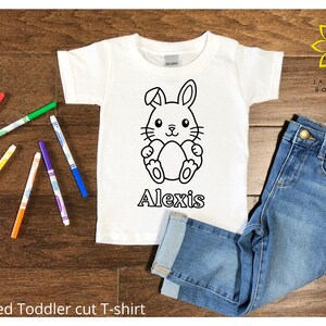 Pode incluir: Camiseta branca para criança pequena com um contorno preto e branco de um coelho segurando um ovo. O nome "Alexis" está impresso abaixo do coelho. A camiseta é mostrada em uma superfície de madeira com marcadores coloridos e calças jeans azuis.