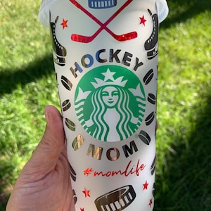 May include: White reusable cup with a black and silver design featuring a hockey stick, puck, and a Starbucks logo with the text "Hockey Mom #momlife".