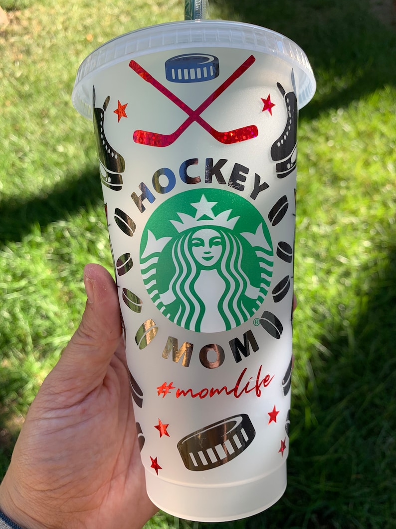 May include: White reusable cup with a black and silver design featuring a hockey stick, puck, and a Starbucks logo with the text "Hockey Mom #momlife".