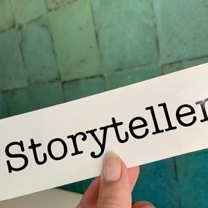 Pode incluir: Cart&atilde;o branco com a palavra "Storyteller" impressa em preto em fonte de m&aacute;quina de escrever.