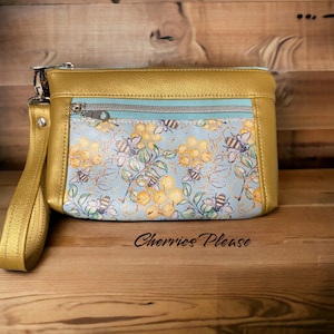 May include: A small gold clutch purse with a zipper closure and a wrist strap. The front of the purse features a blue and yellow pattern of bees and honeycombs. The text "Cherries Please" is printed on the wooden surface below the purse.