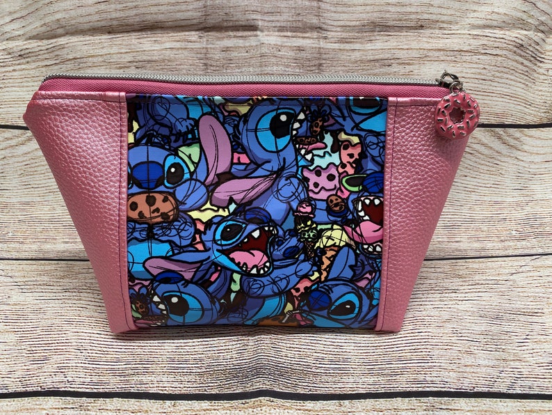 May include: A pink zippered pouch with a colourful print of Stitch characters from the Disney movie Lilo & Stitch. The print features Stitch characters with various expressions and holding different items, including cookies and ice cream cones.