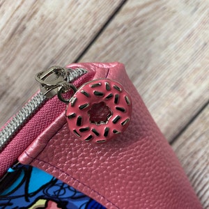 May include: A pink zippered pouch with a donut-shaped zipper pull. The donut is pink with white sprinkles.
