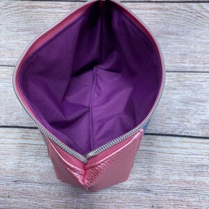 May include: A pink zippered pouch with a purple lining. The pouch is made of a textured material and has a silver zipper.