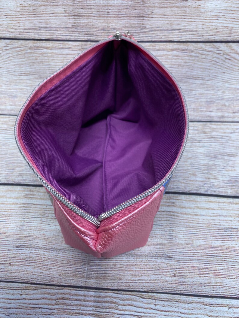 May include: A pink zippered pouch with a purple lining. The pouch is made of a textured material and has a silver zipper.