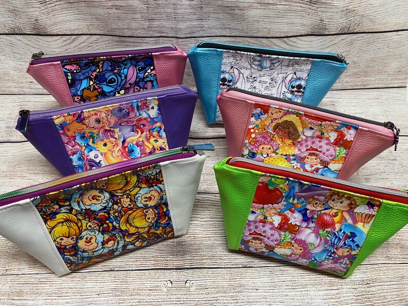 May include: Six colourful zippered pouches with different cartoon character designs. The pouches are made of a faux leather material and have a triangular shape.