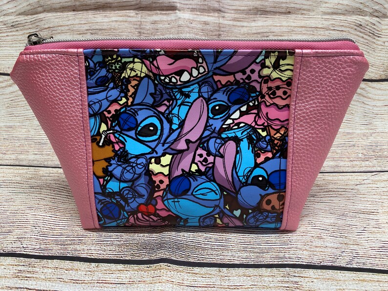 May include: A pink zippered pouch with a colourful print of Stitch characters from the Disney movie Lilo & Stitch. The print features Stitch in various poses and expressions, including a Stitch holding a drink.