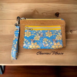 May include: A rectangular clutch with a bee-themed design. The top and sides are brown, with a yellow zipper and a blue front panel featuring bees and honeycomb patterns. A matching blue wrist strap is attached. The text "Cherries Please" is visible.