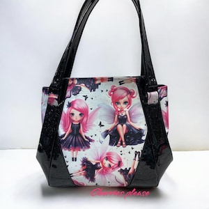 May include: Black tote bag with a glossy finish and black handles. The bag features a print of fairies with pink hair and black dresses on a white background. The text "Cherries please" is printed on the bottom.