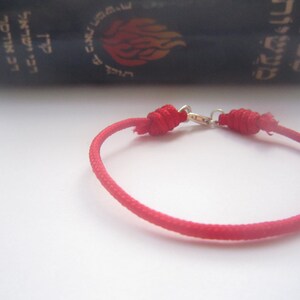 Puede incluir: Un brazalete de cuerda roja con un cierre plateado. El brazalete está atado con un nudo en cada extremo.