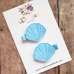 May include: Two light blue felt seashell hair clips with a stitched design. The clips are on a white card with the text "CC's Bows and Beads" and the website address "www.ccsbowandsbeads.com".