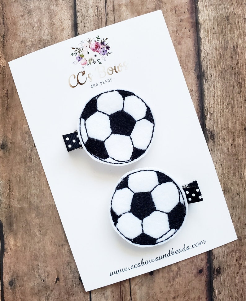 K&ouml;nnte beinhalten: Zwei schwarze und wei&szlig;e Fu&szlig;ball-Haarklammern mit schwarz-wei&szlig;em gepunktetem Band. Die Klammern befinden sich auf einer wei&szlig;en Karte mit dem Text "CC's Bows and Beads" und der Website-Adresse "www.ccsbowandsbeads.com".
