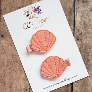 May include: Two coral-colored felt seashell hair clips on a white card with the text "CC's Bows and Beads" and the website address "www.ccsbowsandbeads.com".