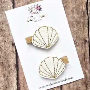 May include: Two white felt seashell hair clips with gold embroidery. The seashells are on a white card with the text "CC's Bows and Beads" and the website address "www.ccsbowandsbeads.com".