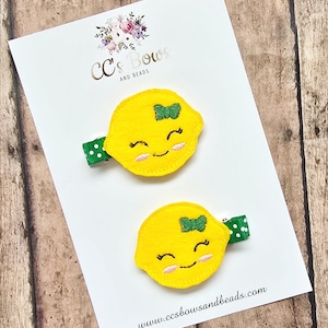May include: Two yellow lemon-shaped hair clips with smiling faces and green bows, attached to green and white polka dot clips. The clips are on a white card with the text "CC's Bows and Beads" and the website address.