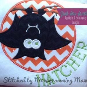 May include: A white t-shirt with an orange and white chevron pattern circle applique featuring a black bat with green eyes. The text "Just Sew Sweetly Applique & Embroidery Designs" is in a purple banner above the circle. The text "Stitched by Monogramming Mamacita" is below the circle. The text "HATCHER" is in green text on the right side of the shirt.