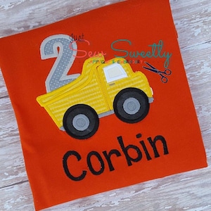 May include: A red t-shirt with a yellow dump truck, a gray number 2, and the name Corbin in black embroidery. The text "Just Sew Sweetly" is also embroidered on the shirt.